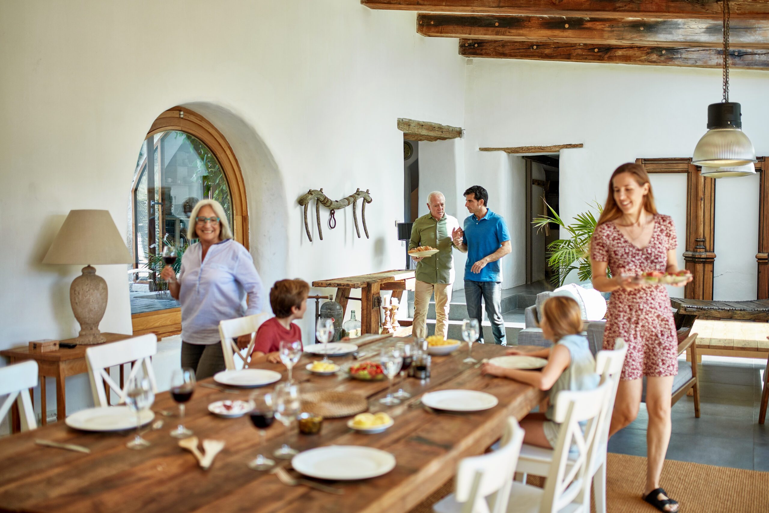 Adults approaching Spanish-style farmhouse dining table with food and drink as seated children wait for start of lunch.